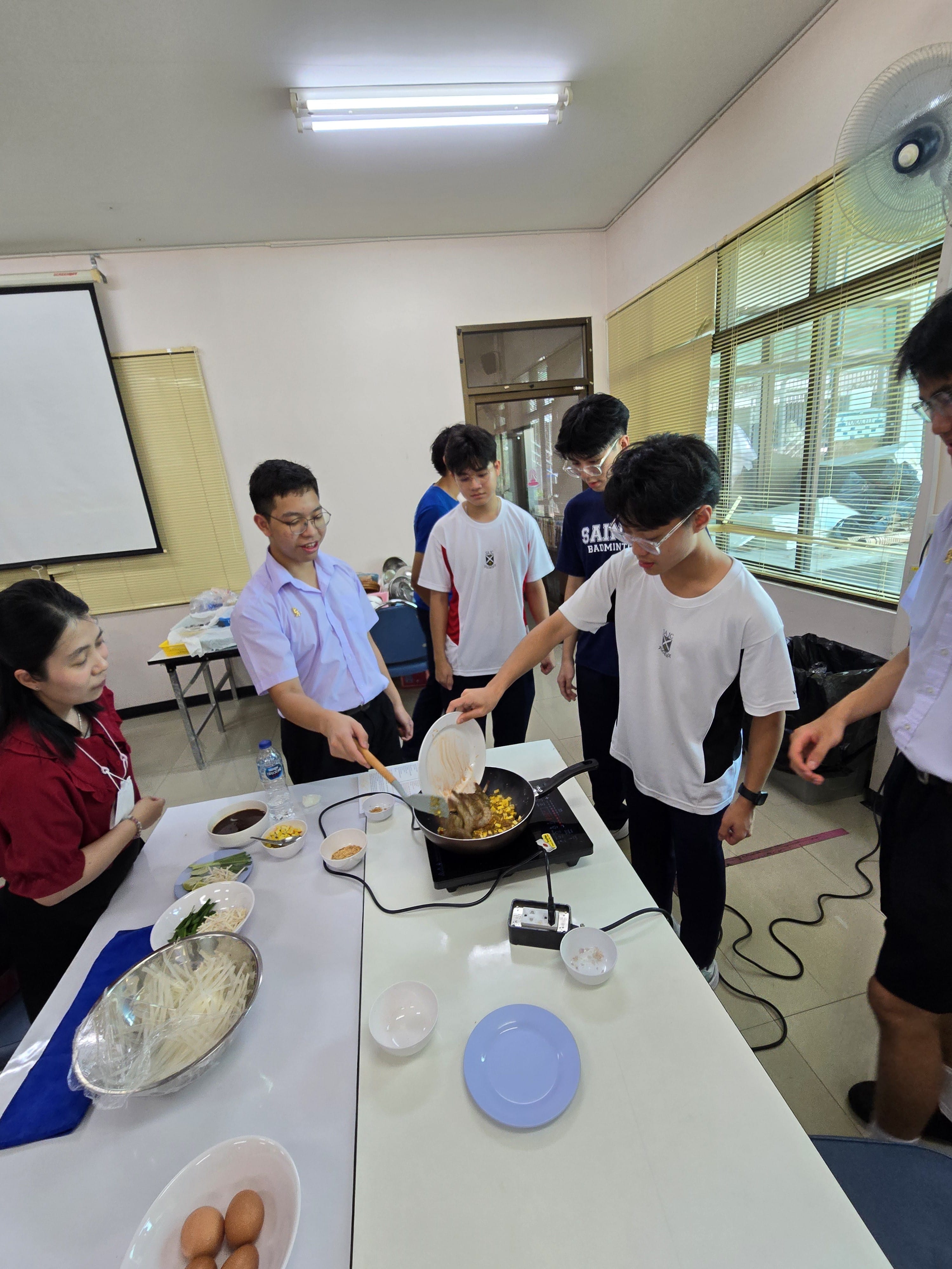 Students gathered around a table watch as a Thai student in school uniform demonstrates stir-frying in a wok on an induction cooker, with various ingredients and plates laid out on the table.